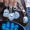 A hand holds a clear ghost-shaped glow-in-the-dark keychain featuring cute, smiling ghosts against a brown leather background