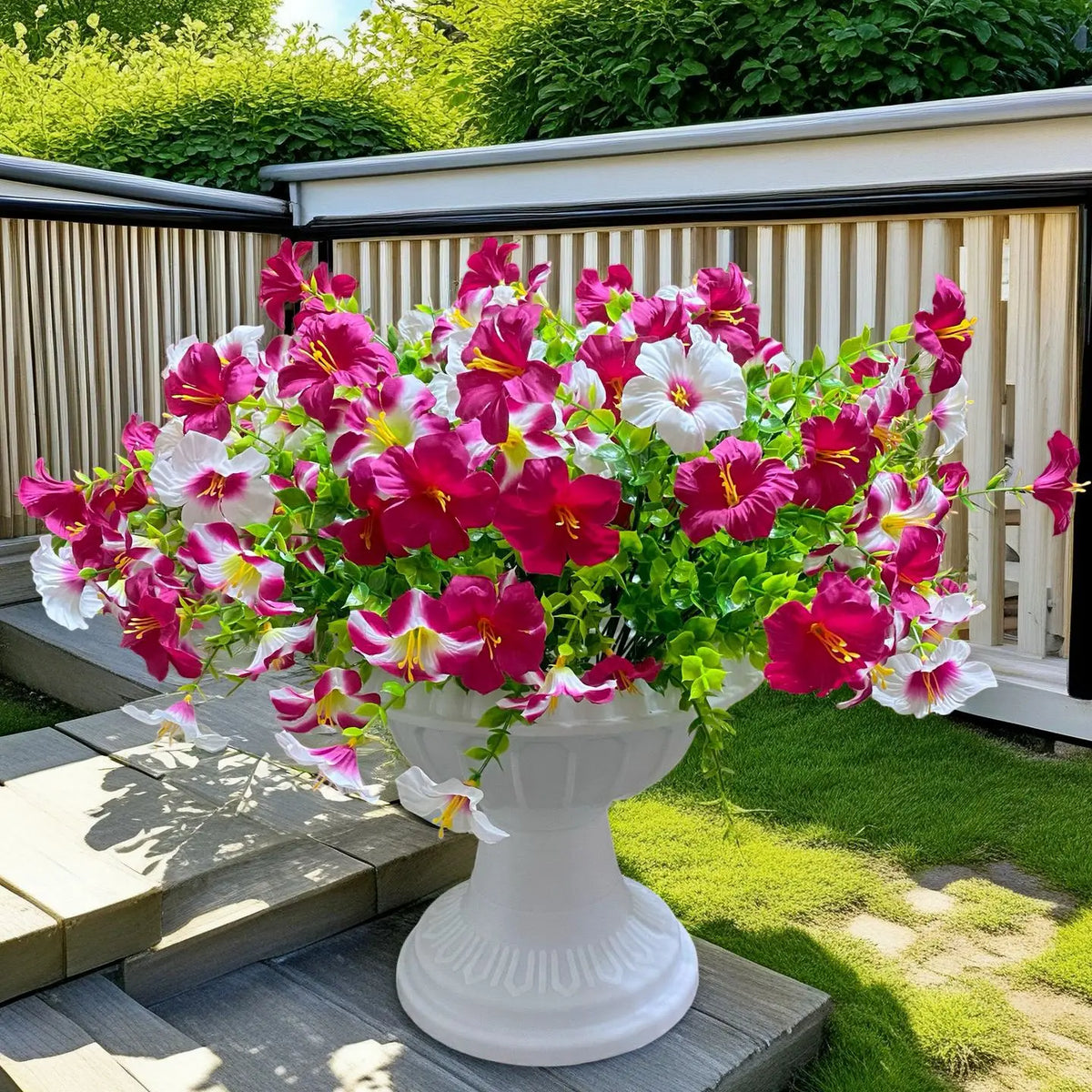 A vibrant bouquet of artificial silk pink and white flowers in a white pedestal vase set on an outdoor deck.