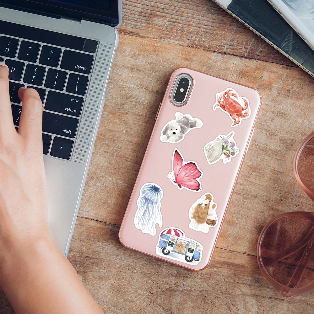 A pink smartphone displays a watercolor sticker pack featuring various animals and objects on a wooden table next to a laptop