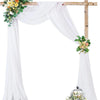 A white chiffon wedding arch with draping fabric panels and a wooden cross, decorated with flowers.