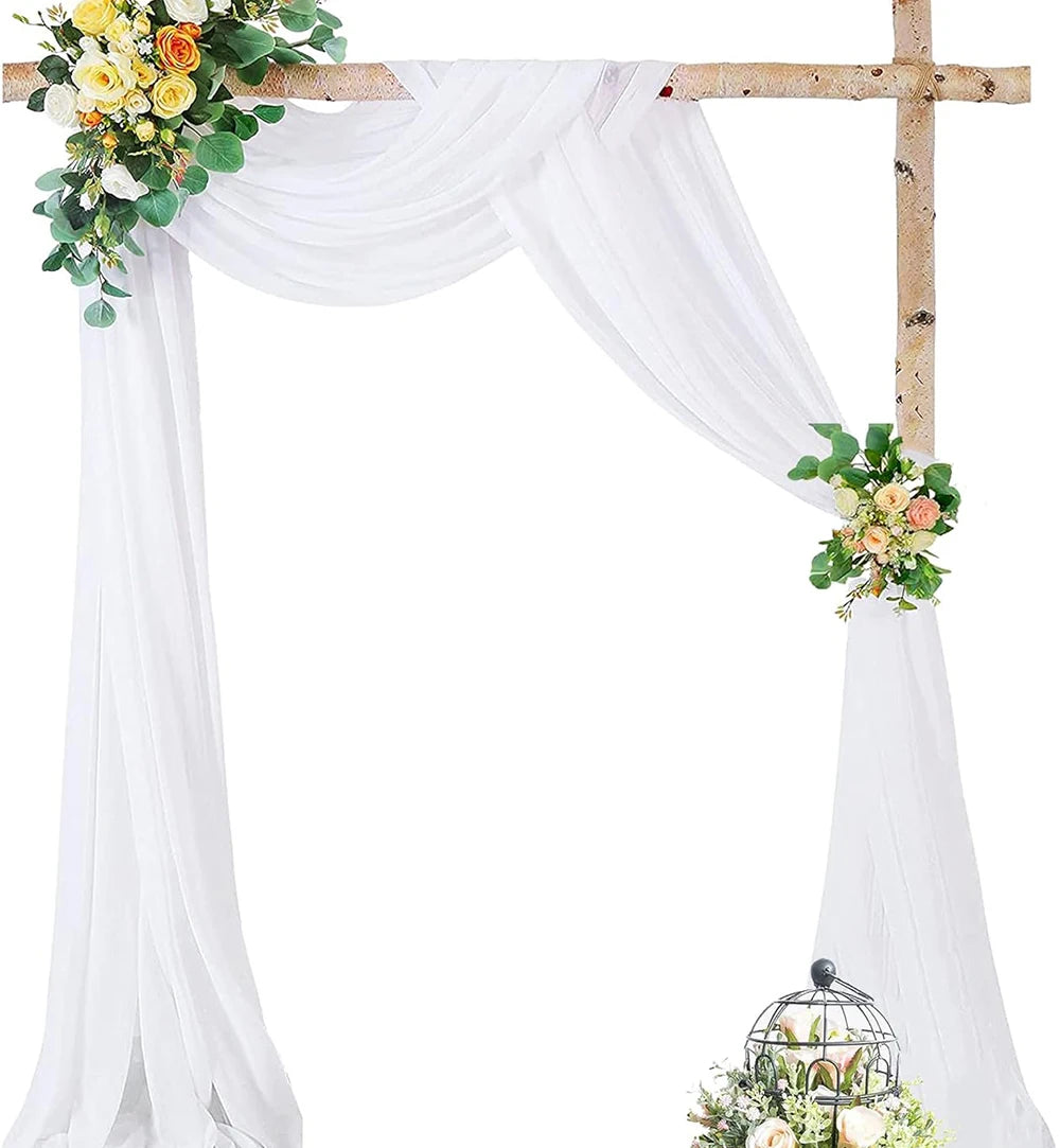 A white chiffon wedding arch with draping fabric panels and a wooden cross, decorated with flowers.