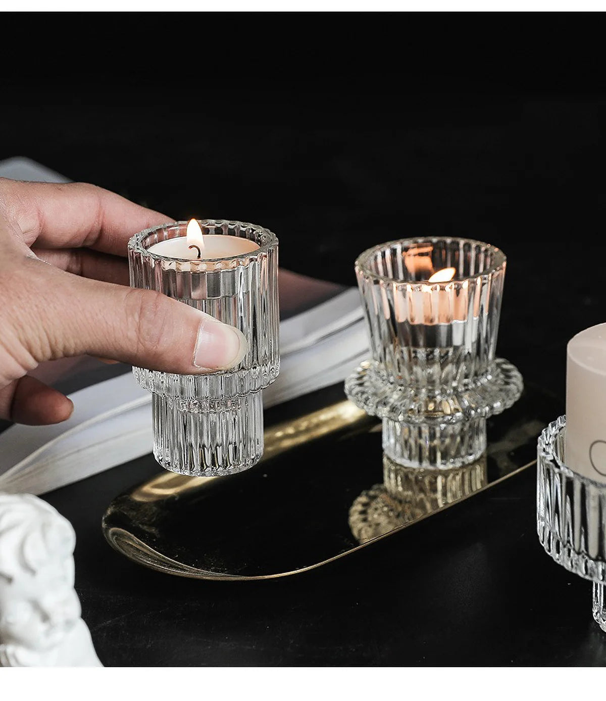 Una mano sostiene un portavelas de cristal transparente con una vela encendida sobre una bandeja dorada.