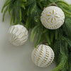 A set of retro shatterproof Christmas tree ball ornaments with white and gold patterns hanging on green pine branches.