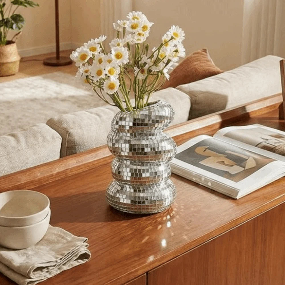 A silver disco ball spiral vase holding white flowers on a wooden desk with a book and a cloth.