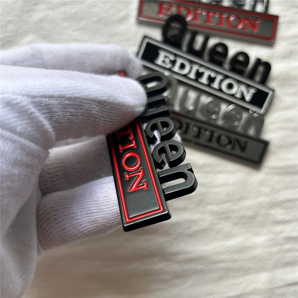 Eine Hand in weißer Handschuh hält ein Set aus schwarzen Emblem-Autoklebern mit der Aufschrift Queen Edition