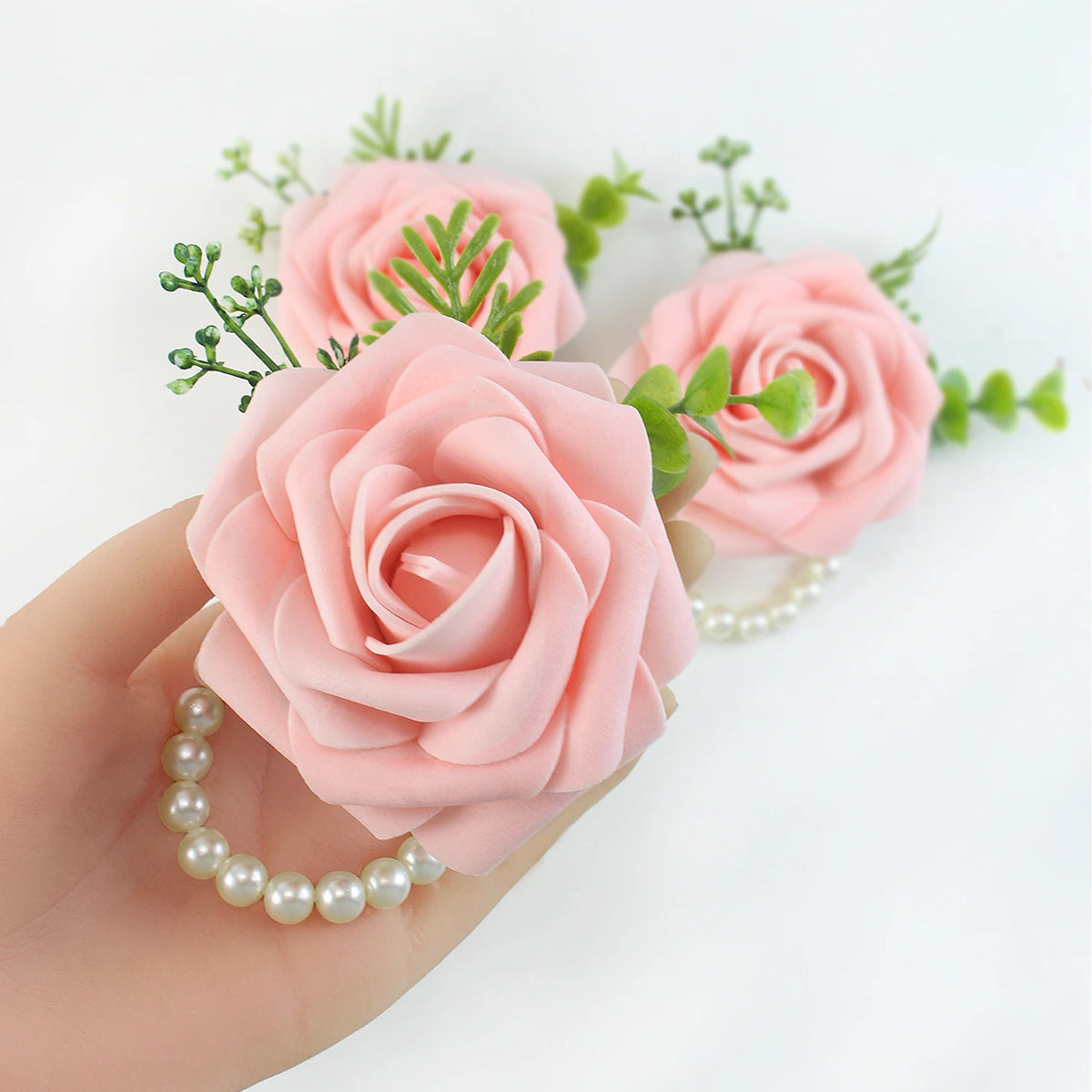 A hand holds an elegant wrist corsage bracelet featuring pink silk roses and green foliage.
