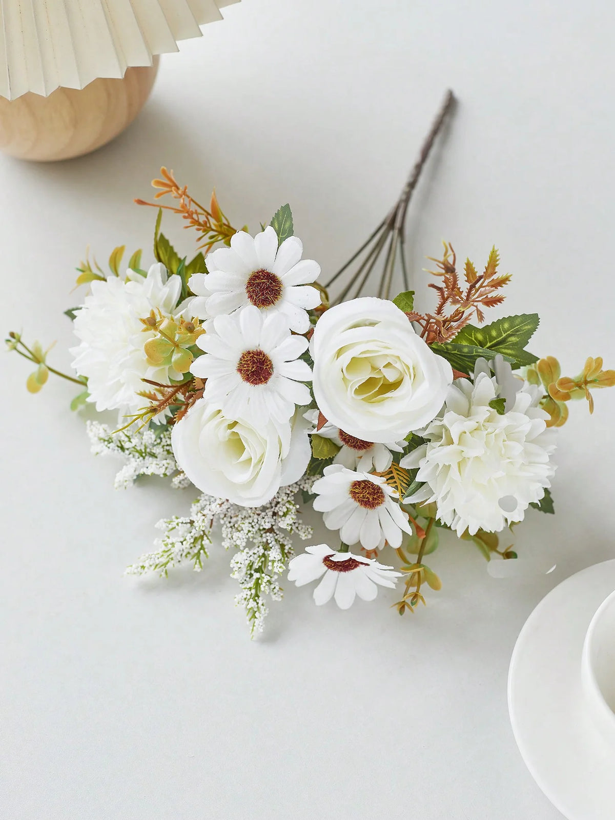 A bouquet of artificial white and cream silk flowers with green foliage and dried accents on a white surface.