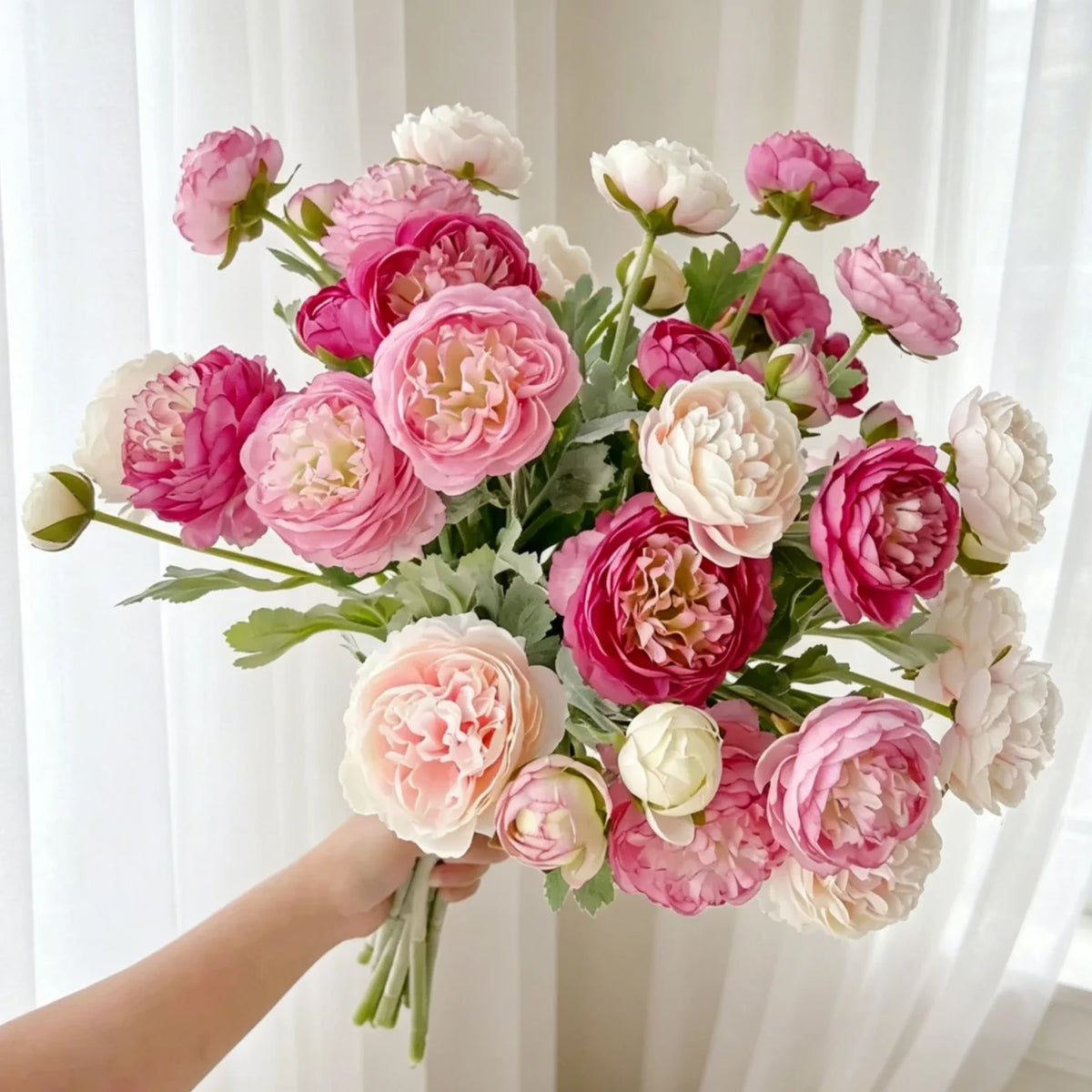 A hand holds a bouquet of lifelike silk peony flowers in shades of pink and white.