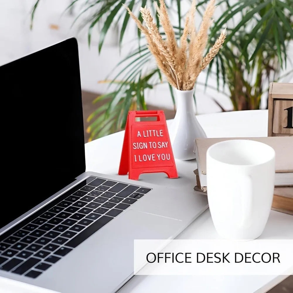 A red 3D printed letter sign saying I love you is placed on a white office desk next to a laptop and greenery.