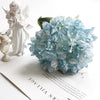 A light blue silk hydrangea flower bouquet with vintage burnt edge petals next to a white angel figurine.