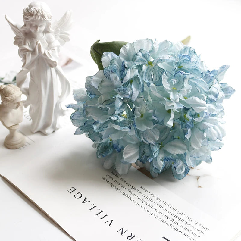 A light blue silk hydrangea flower bouquet with vintage burnt edge petals next to a white angel figurine.