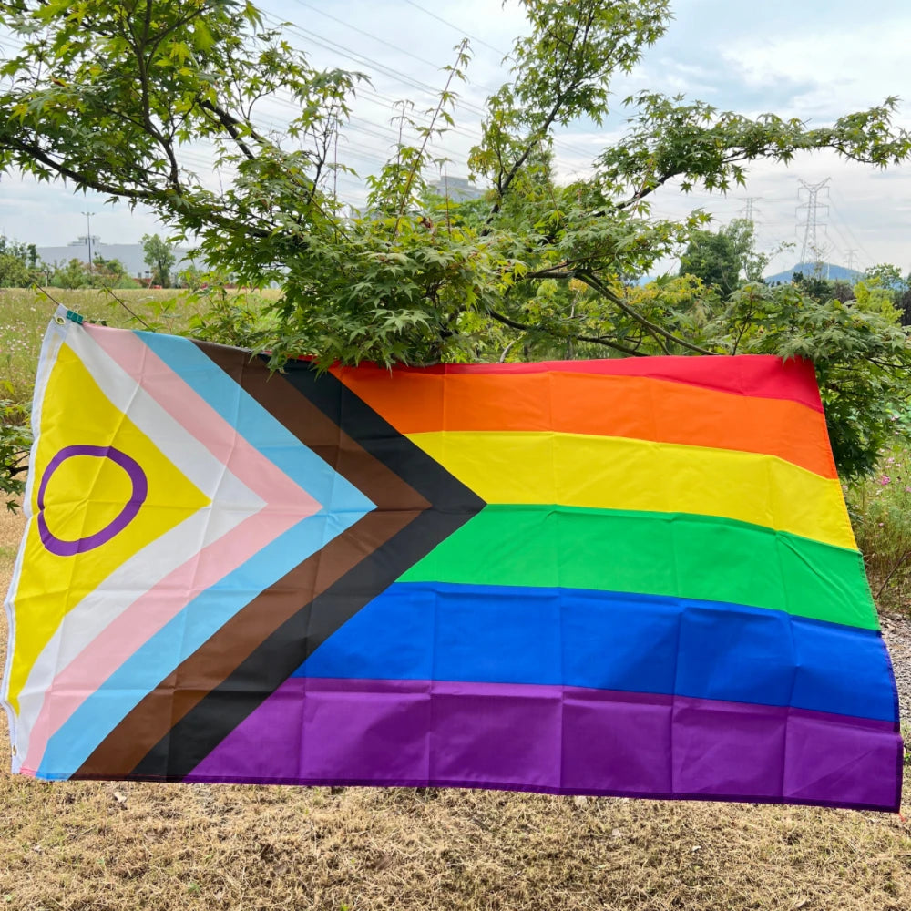 A large, colorful Pride rainbow flag banner is displayed outdoors against a backdrop of green trees and a hazy sky.