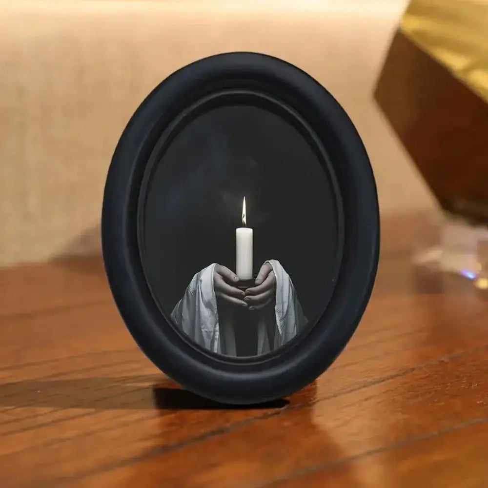 A black candle is held by hands within a dark, oval frame, set on a wooden surface.