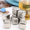 A pile of reusable stainless steel ice cubes resting on a white surface next to a glass of amber liquid.