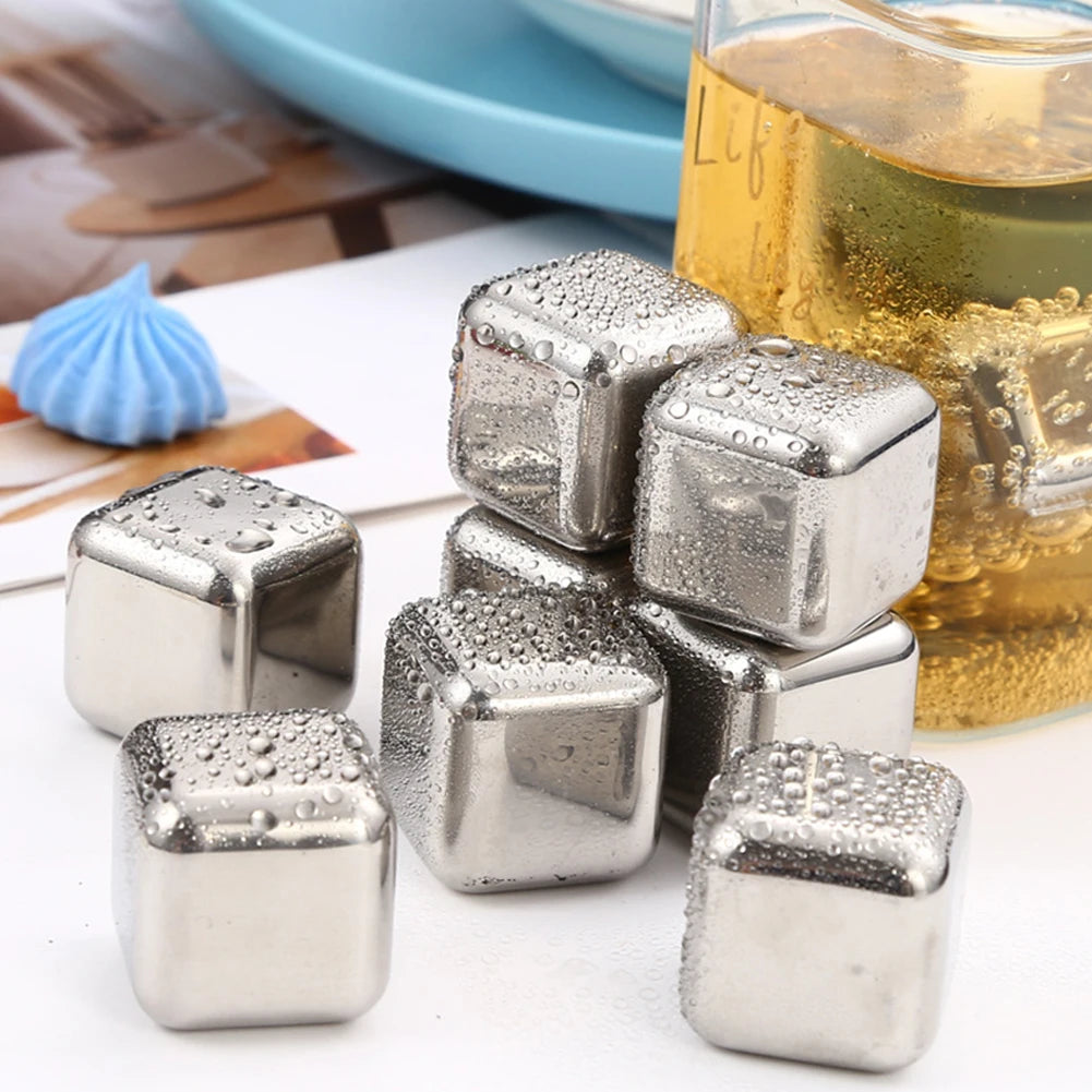A pile of reusable stainless steel ice cubes resting on a white surface next to a glass of amber liquid.