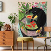 A tapestry featuring a black cat and goldfish in a glass bowl, displayed on a wall above a wooden cabinet.