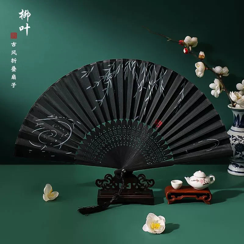 A black vintage silk bamboo folding hand fan displayed against a dark green background with small white flowers.