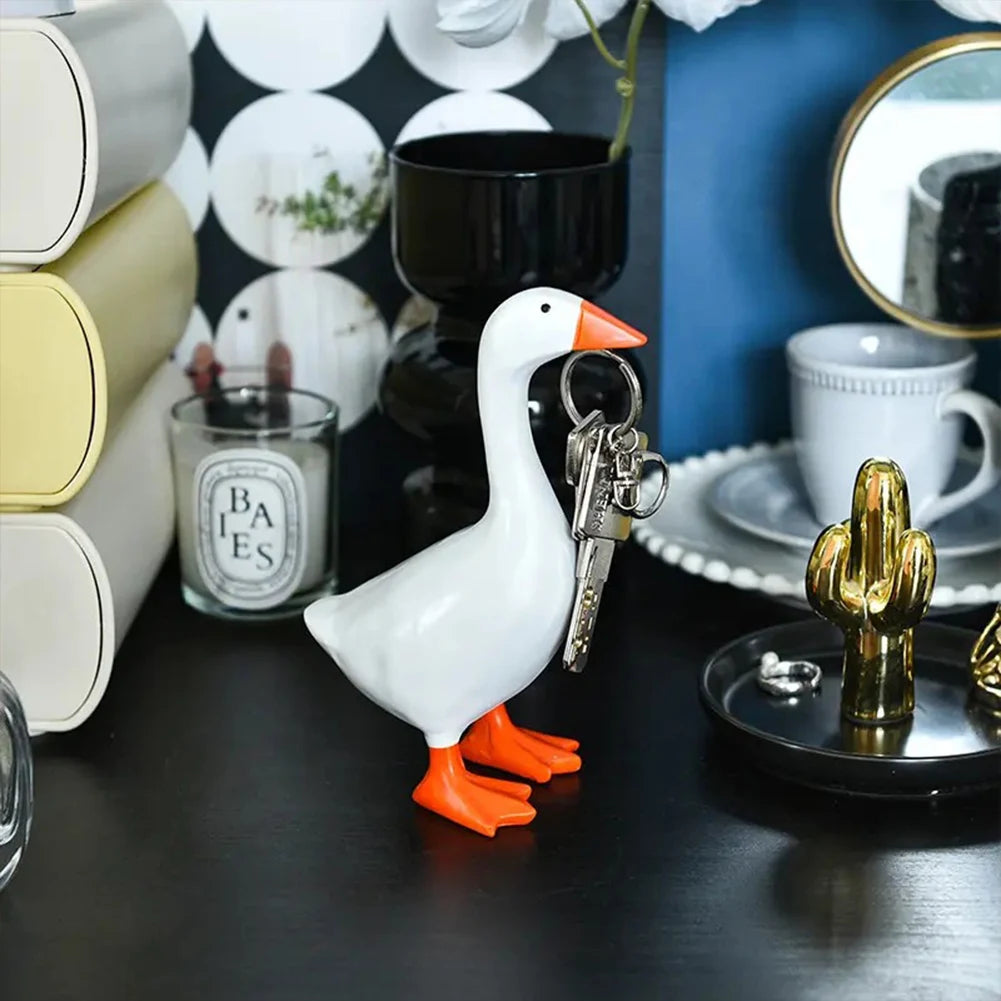 A white ceramic goose figurine with orange feet and a silver key attached, displayed on a dark surface.