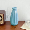 A light blue ceramic bud vase is shown on a wooden surface next to a vintage-style radio.