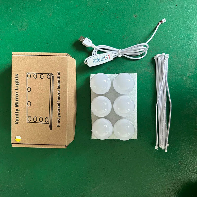 A box containing a dimmable LED vanity mirror, its power cord, and several round light bulbs on a green surface.