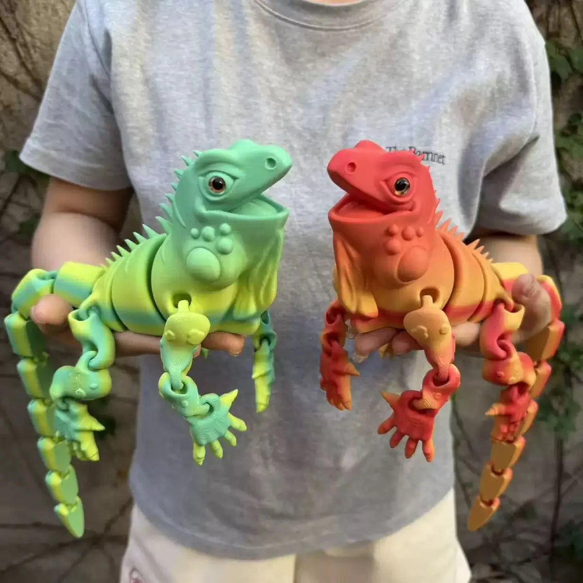 A person holds a bright green articulated 3D printed lizard figurine and an orange lizard figurine.