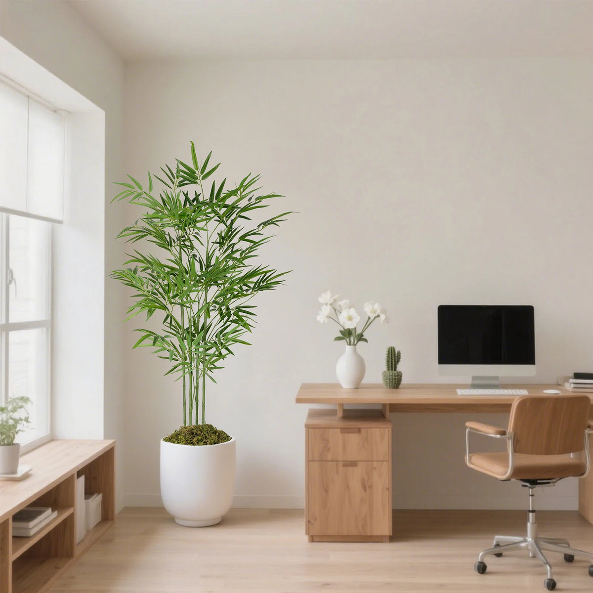 A tall artificial bamboo tree in a white pot sits on a wooden desk in a bright room.