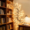 A decorative branch of flexible willow LED lights illuminates a wooden bookshelf and desk area.