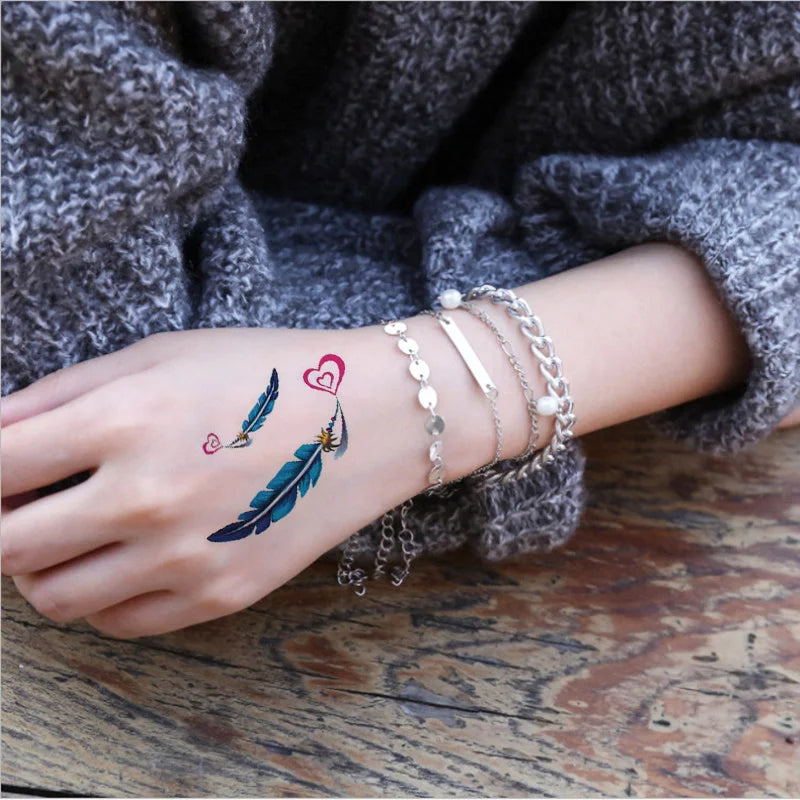 A hand wearing silver bracelets and a temporary body art tattoo featuring a bird feather and a heart on the wrist.