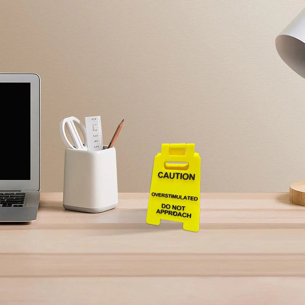 A yellow caution desk sign with text warning against approaching due to overstimulation sits on a wooden desk.