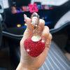 A hand holds a red heart-shaped crystal keychain with a gold chain and charm against a blurred background.