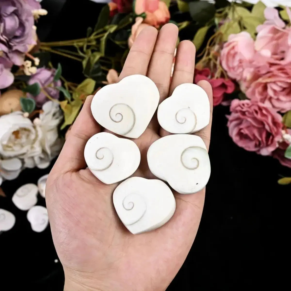 A hand holds several white rose quartz heart-shaped stones with swirling designs against a backdrop of pink and purple roses.