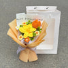 A bouquet of handmade crochet flowers in yellow and orange sits inside a light brown paper bag.