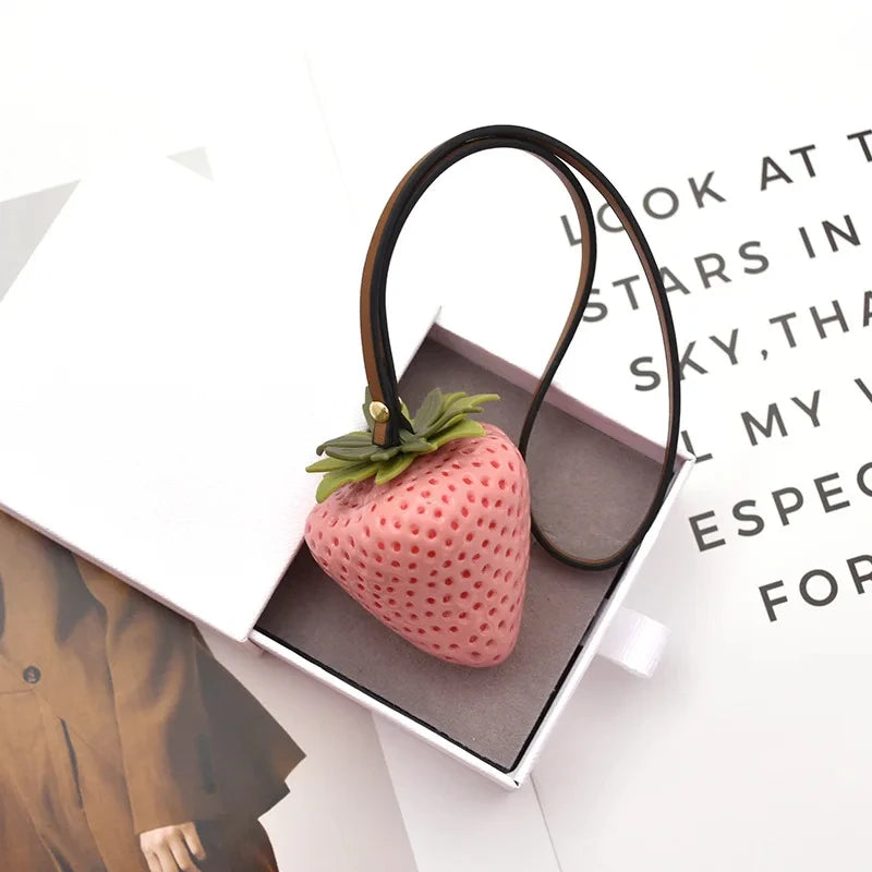A pink strawberry charm and leather cord keychain displayed in a box.