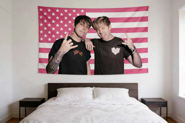 Two young men pose in front of a patriotic American flag tapestry hanging above a bed.
