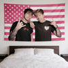 Two young men pose in front of a patriotic American flag tapestry hanging above a bed.