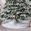 A green artificial Christmas tree decorated with silver and gold ornaments and a white snowflake-patterned skirt.