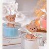 A soy wax teddy bear candle in a glass jar with a ribbon, set on a white surface.