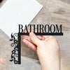A hand holds up a black metal wall plaque shaped like the word BATHROOM with floral accents.