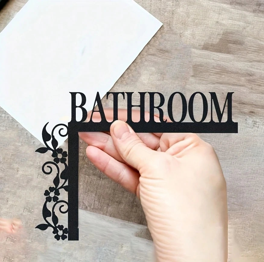 A hand holds up a black metal wall plaque shaped like the word BATHROOM with floral accents.