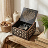 An antique wood music box with an engraved melody sits on a wooden surface next to a potted plant and books.