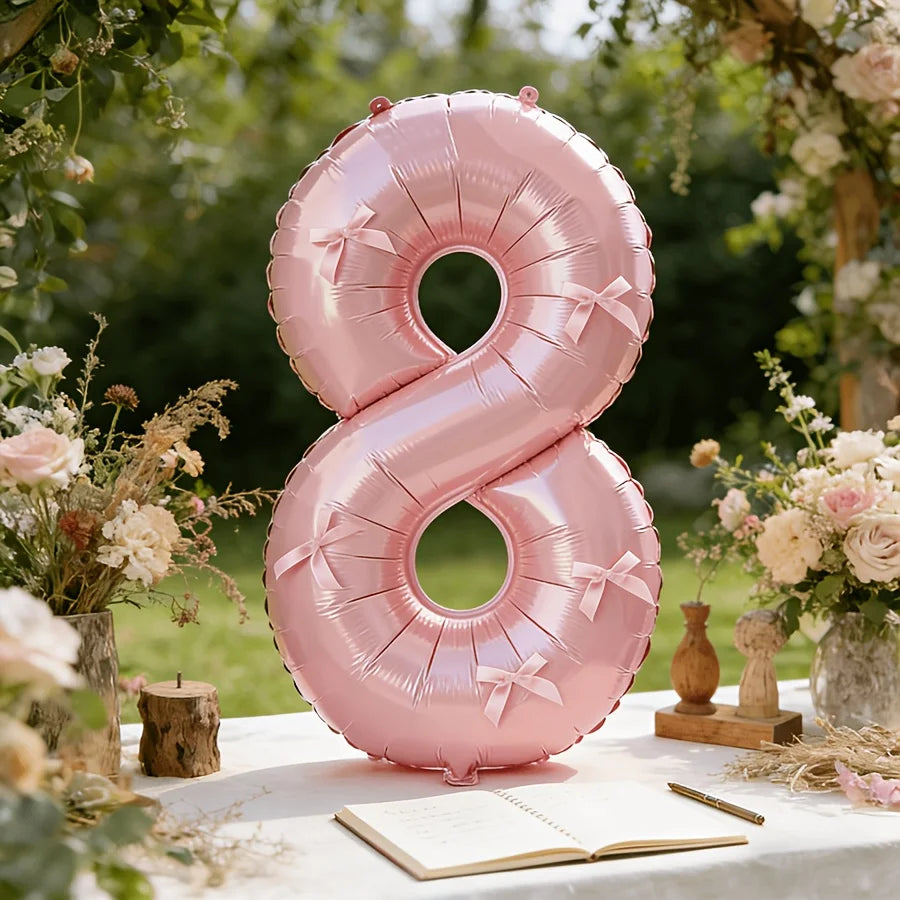 A large pink foil number balloon shaped like the number eight with bows, displayed outdoors among greenery and flowers.