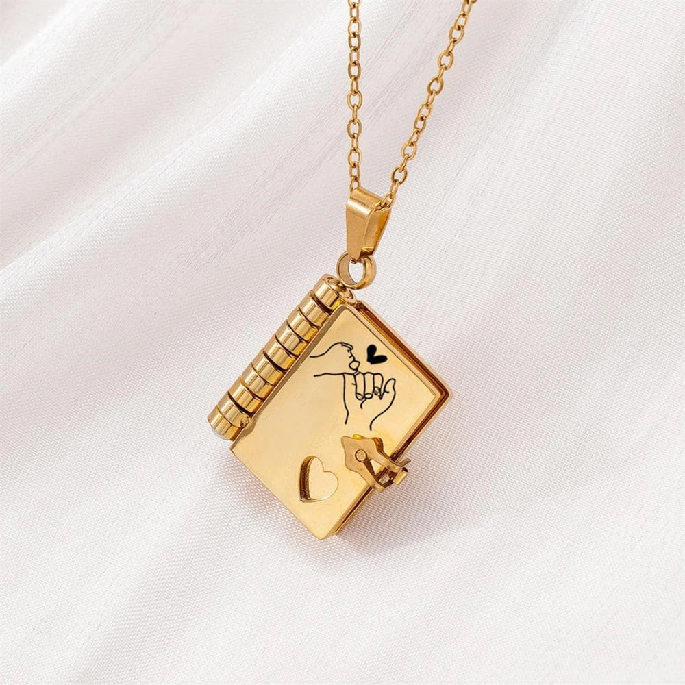 A gold-colored openable book pendant necklace with an engraved hand heart design displayed on white fabric.