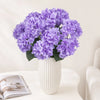 A bouquet of purple artificial hydrangea silk flowers in a white vase on a light surface.