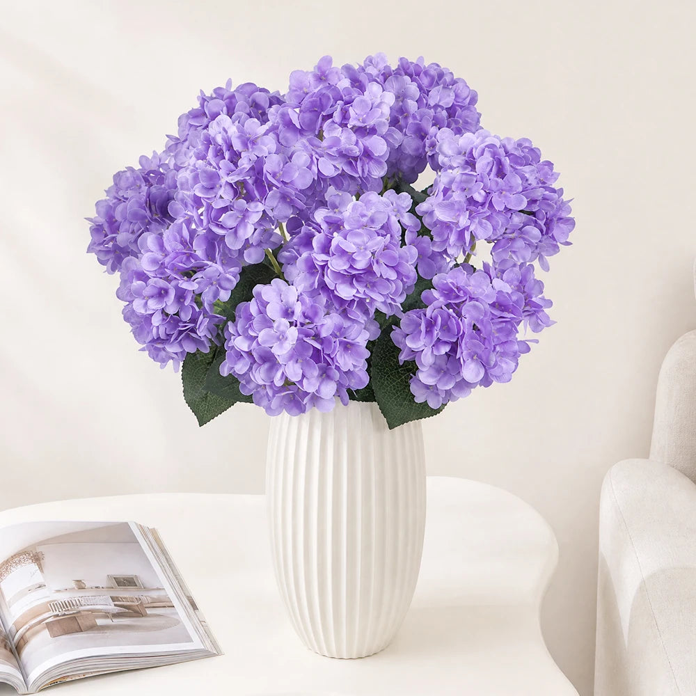 A bouquet of purple artificial hydrangea silk flowers in a white vase on a light surface.