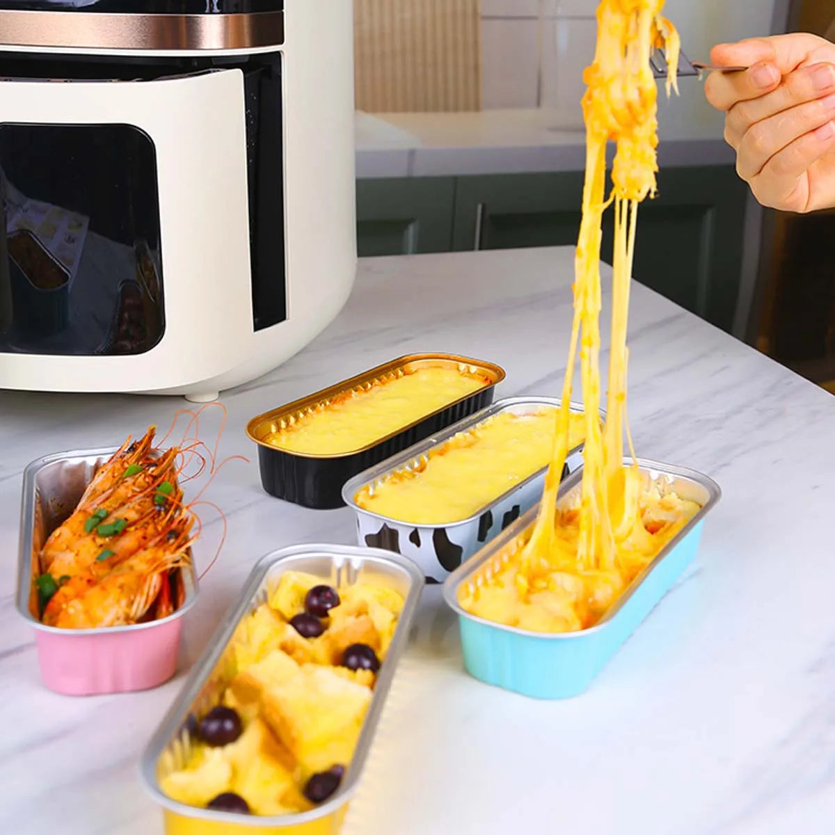 A hand is lifting strands of yellow food from a mini foil bread tray container on a white surface.