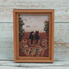 A framed painting featuring two black cats silhouetted against a sunset and foliage.