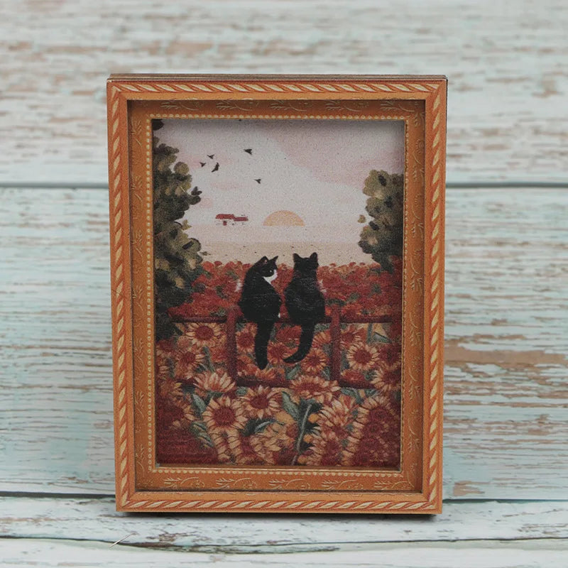A framed painting featuring two black cats silhouetted against a sunset and foliage.