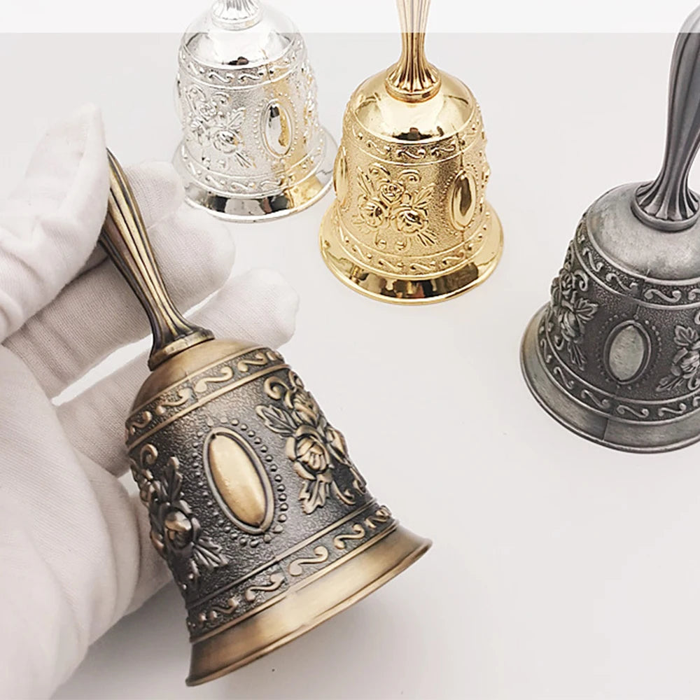 Three ornate brass hand call bells in silver, gold, and dark metal resting on a white surface.