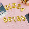 Gold acrylic number cake candles shaped like 1, 2, 3, 4, 5, 6, 7, 8, and 9 arranged on a pink background.
