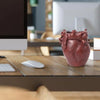 A reddish-brown heart-shaped resin pen organizer sits on a wooden desk next to a computer monitor and a white mouse.